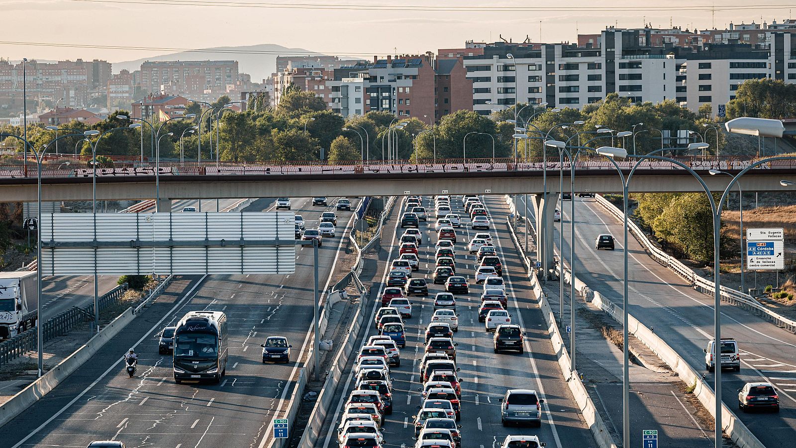 Vista aérea de autopista A-3 con tráfico denso al atardecer/amanecer, señal "P.I. Arganda", puente y entorno urbano.