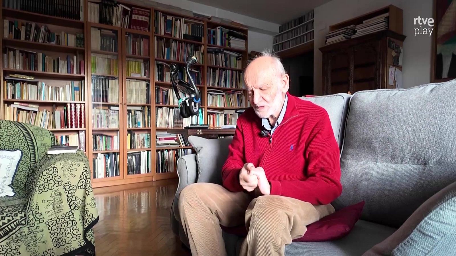 Antonio Gamoneda, poeta español, en un salón con estanterías de libros. Viste jersey rojo y pantalón beige. Logo de "rtve play" visible.