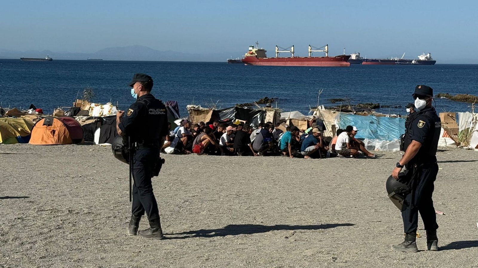 Agentes de la Policía Nacional en la Playa del Trampolín de Ceuta, junto a un campamento de migrantes y el mar Mediterráneo al fondo.
