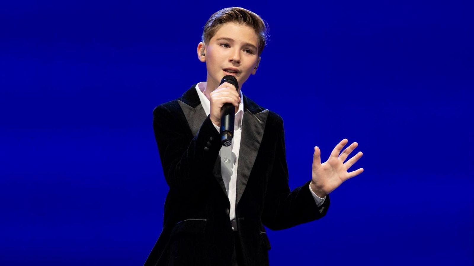 Primer plano de Lucas Paulano cantando, con chaqueta de terciopelo negro, micrófono en mano y gesto expresivo. Fondo azul sólido.