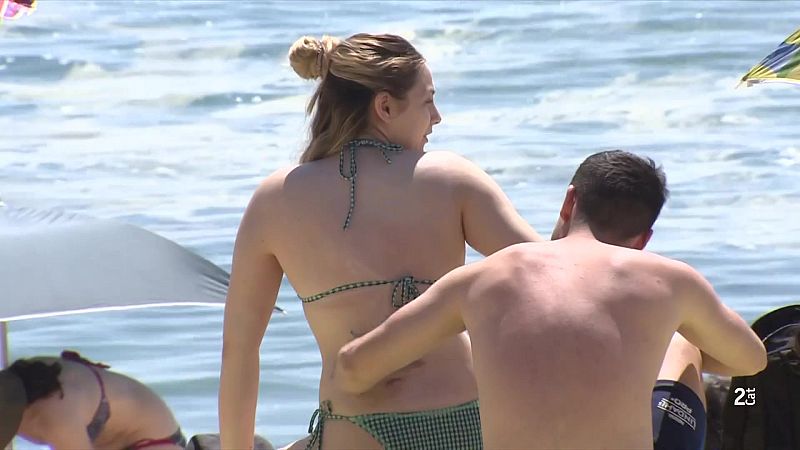 Playa soleada con pareja: mujer en bikini de cuadros verdes y blancos, hombre en bañador azul oscuro. Temperatura de 23 grados.