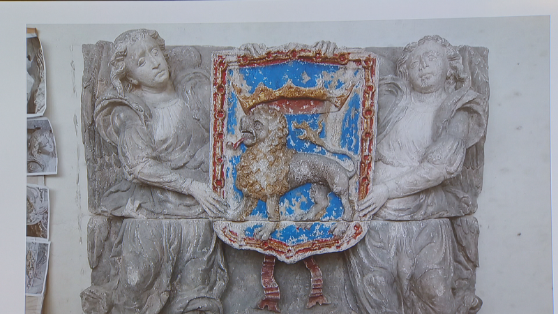 Escultura en relieve del escudo de Pamplona con león rampante coronado, cadenas rojas y figuras alegóricas sosteniéndolo. Realizada en piedra policromada y grisácea.