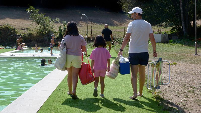 Familia se dirige a piscina municipal en día soleado. Mujer con bolsos, hombre con nevera y silla, niña de rosa. Césped artificial y agua verde.