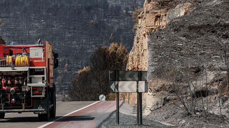 Camión de bomberos rojo con "PROTECC. FI" parcialmente visible, sobre carretera gris. Fondo de montaña calcinada y señal de peligro. Paisaje desolador post-incendio.