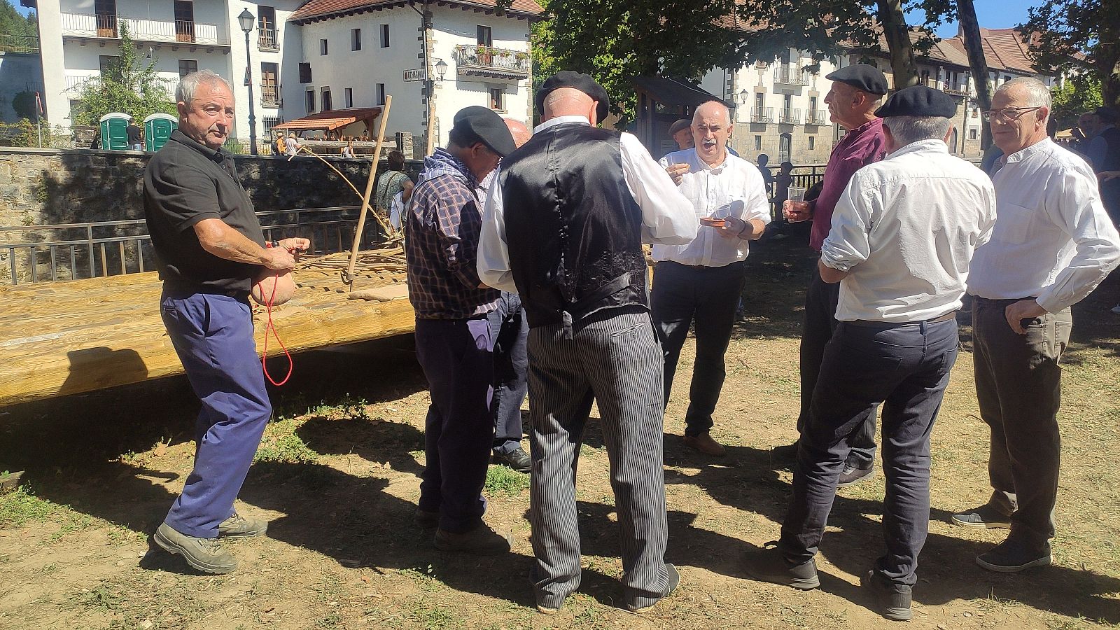 Hombres de edad avanzada, algunos con boinas, participan en un evento al aire libre. Uno sostiene una cuerda atada a un objeto marrón. Al fondo, edificios tradicionales y cabinas verdes.