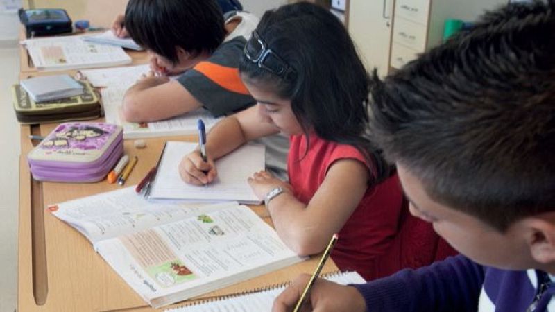 Tres niños estudian en un aula. Uno escribe en primer plano, una niña de perfil con diadema y reloj escribe en el centro, y otro niño se inclina al fondo. Pupitres de madera clara y mobiliario escolar completan la escena.