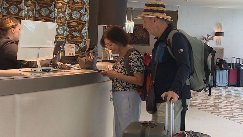 En una recepción de hotel, una recepcionista atiende a dos viajeros. Uno consulta su móvil, el otro sujeta una maleta. El fondo presenta decoración y maletas apiladas.