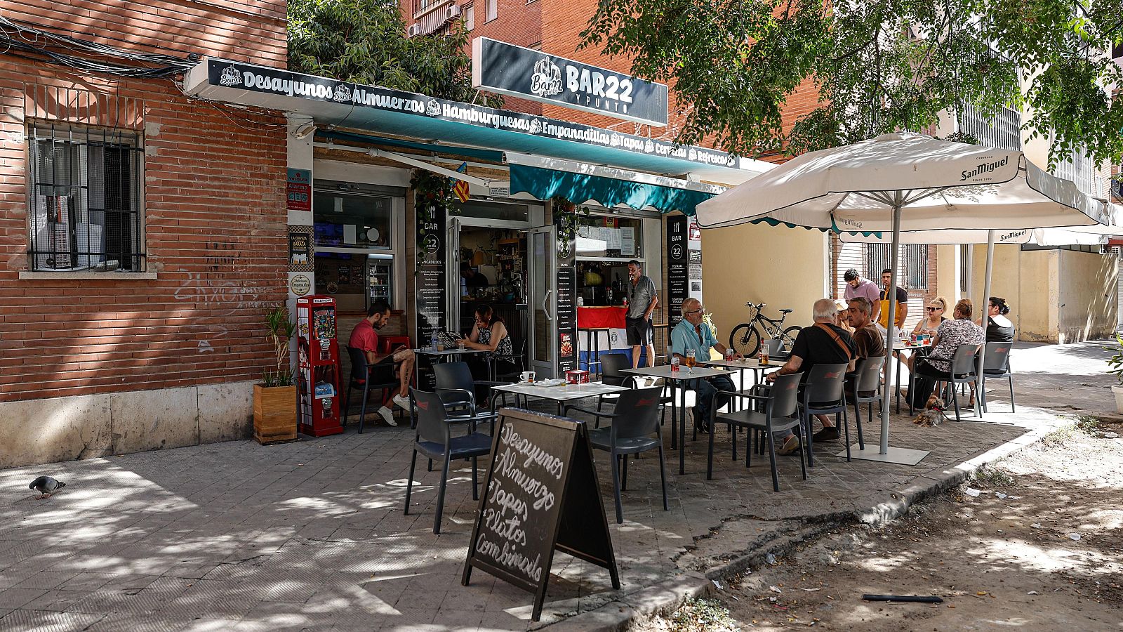 Fachada de "BAR22 Y TAPAS" con terraza soleada. Toldo con menú: "Desayunos Almuerzos Hamburguesas Empanadillas Tapas Cerveza". Pizarra: "Desayunos Almuerzos Tapas Platos Combinados".