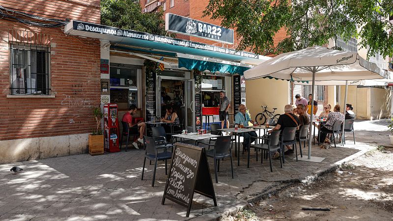 Fachada de "BAR22 Y TAPAS" con terraza soleada. Toldo con menú: "Desayunos Almuerzos Hamburguesas Empanadillas Tapas Cerveza". Pizarra: "Desayunos Almuerzos Tapas Platos Combinados".