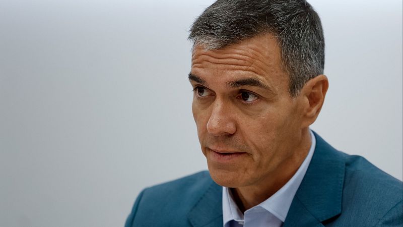 Primer plano de Pedro Sánchez, Presidente del Gobierno de España, con expresión seria. Viste traje azul oscuro y camisa azul claro. Fondo liso y claro.