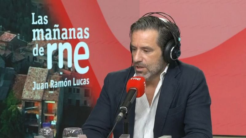 Juan Ramón Lucas en el estudio de "Las mañanas de RNE", con auriculares y micrófono, vestido con americana azul y camisa blanca. Fondo rojo con gráfico del programa.