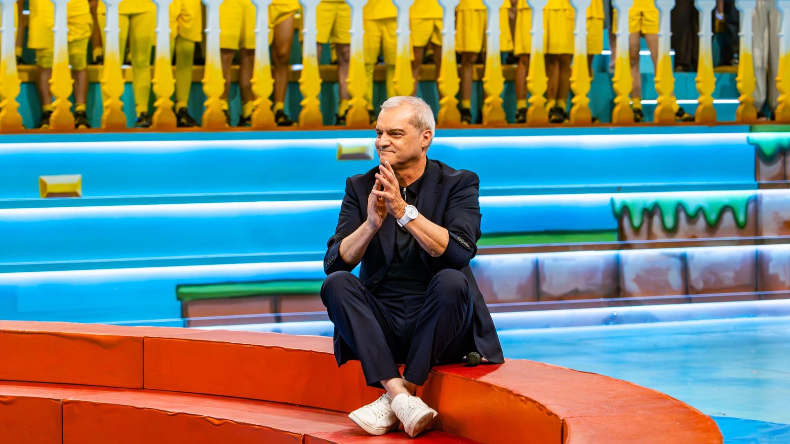 Ramón García, presentador, sentado en un plató de TV azul y naranja con decoración de videojuegos, durante la final del "Grand Prix del Verano 2026".
