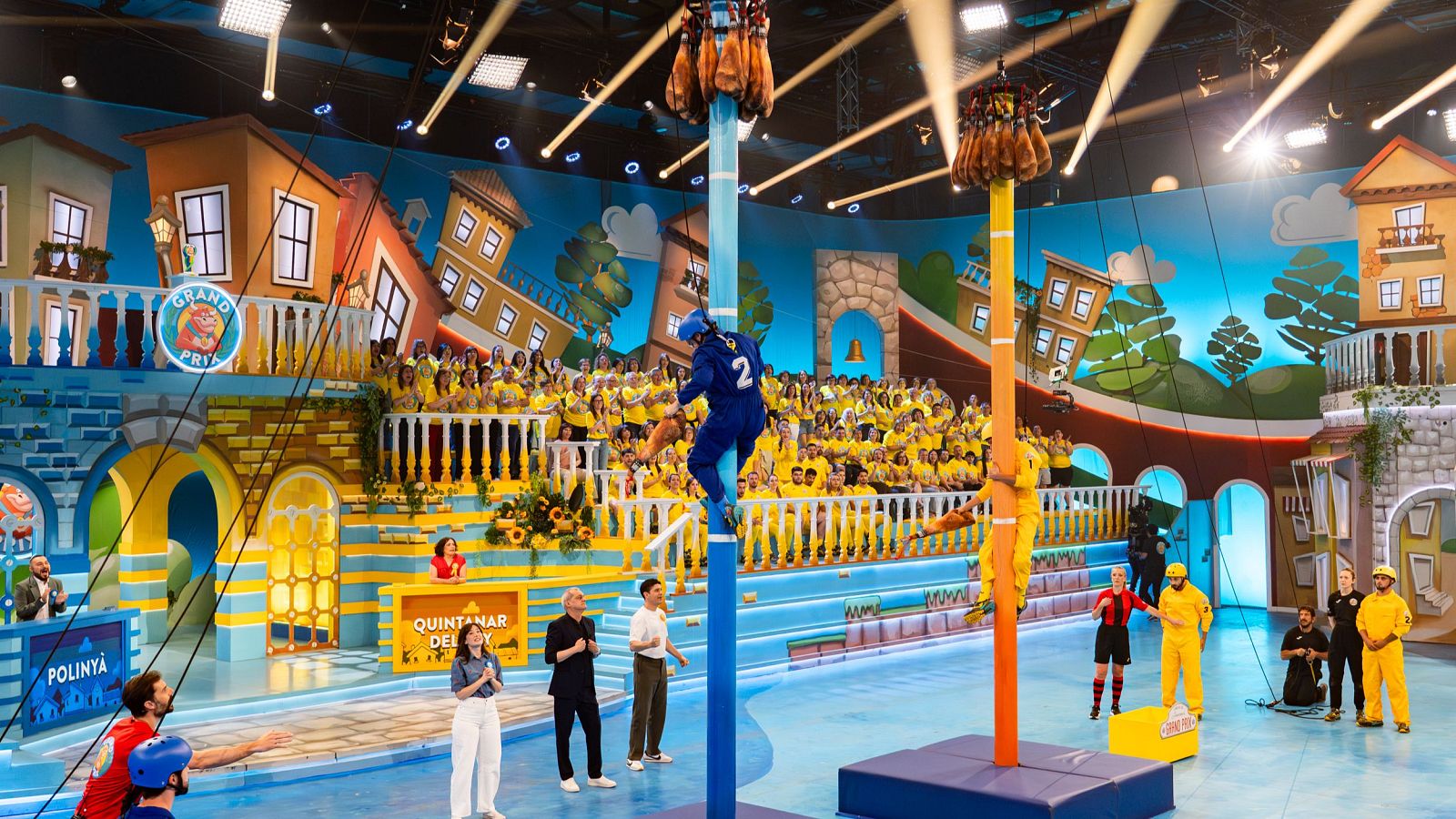 Concursantes de Polinyà y Quintanar del Rey escalan postes en "La Cucaña" durante la final del "Grand Prix del Verano", ante un público entusiasta y la atenta mirada de Ramón García y Michelle Calvó.