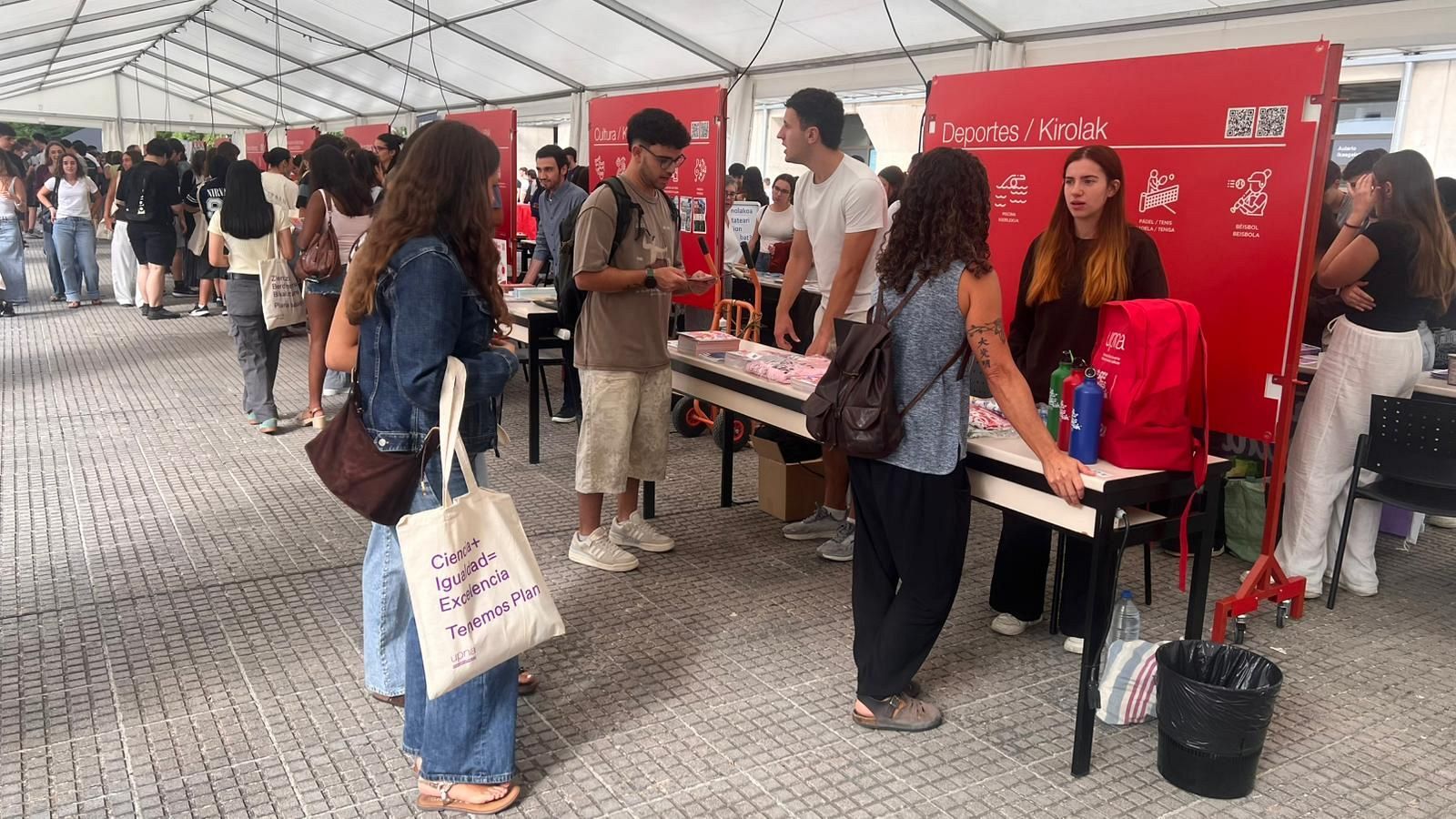 Evento bajo carpa blanca con stand "Deportes / Kirolak". Jóvenes interactúan en puestos informativos. Mensaje "Crea+ Igualdad= Excelencia Tejemos Plan" visible en un bolso.