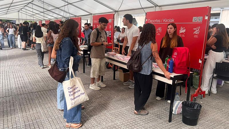 Evento bajo carpa blanca con stand "Deportes / Kirolak". Jóvenes interactúan en puestos informativos. Mensaje "Crea+ Igualdad= Excelencia Tejemos Plan" visible en un bolso.