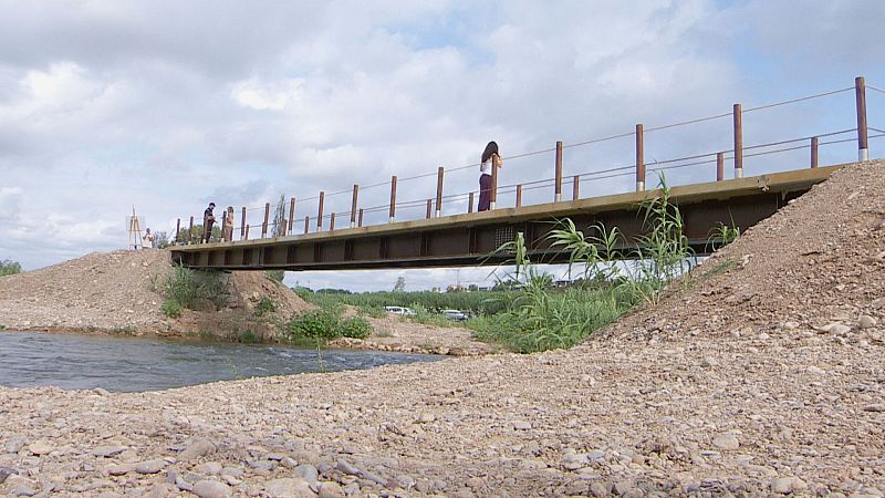 Pasarela peatonal de metal y madera sobre un río, con personas transitando y un topógrafo en la orilla. Proyecto de infraestructura en curso.