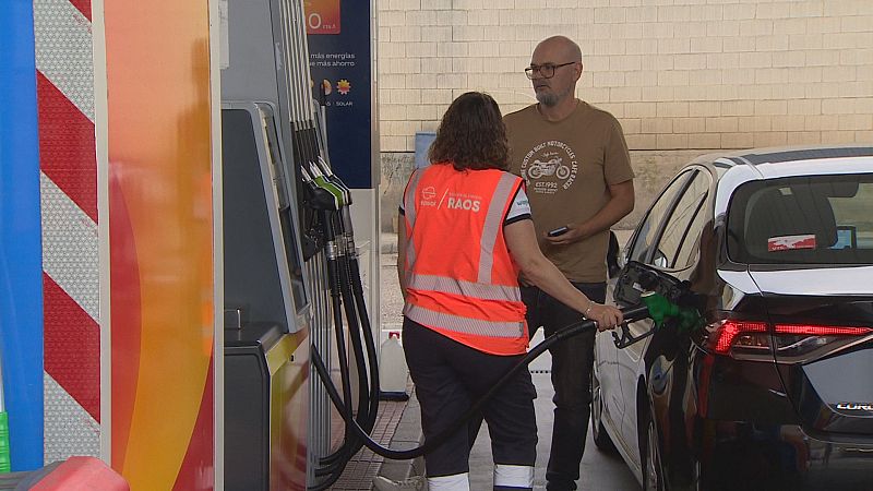 En una gasolinera, una empleada con chaleco "RAOS" reposta un Toyota Corolla. Un cliente con camiseta "CUSTOM BUILT MOTORCYCLES" mira su móvil. Cartel: "más energías más ahorro".