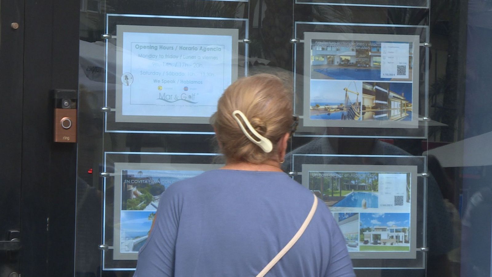 Mujer rubia/castaña observa escaparate de agencia inmobiliaria "Mar & Golf" con anuncios de propiedades y horario.