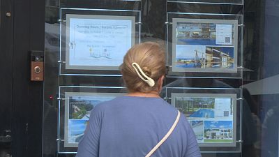 Mujer rubia/casta�a observa escaparate de agencia inmobiliaria "Mar & Golf" con anuncios de propiedades y horario.