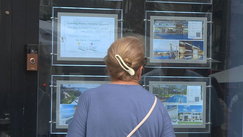 Mujer rubia/castaña observa escaparate de agencia inmobiliaria "Mar & Golf" con anuncios de propiedades y horario.