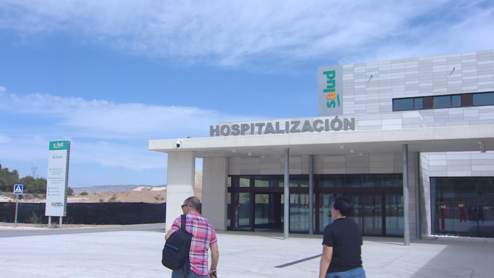 Entrada de un hospital moderno con señalización "HOSPITALIZACIÓN" y "Salud". Dos personas se dirigen hacia el edificio. Cielo despejado con nubes.