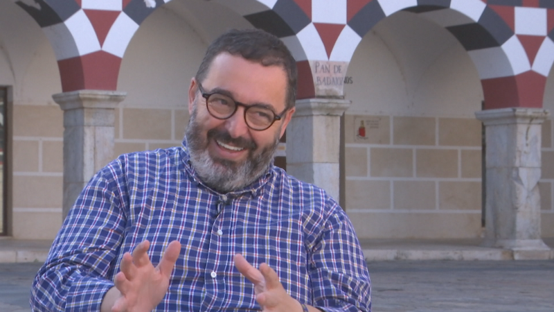 Núñez Medalla, con barba canosa y gafas, sonríe y gesticula. Detrás, arcos decorados con damero y cartel "PAN DE BADAJOZ".