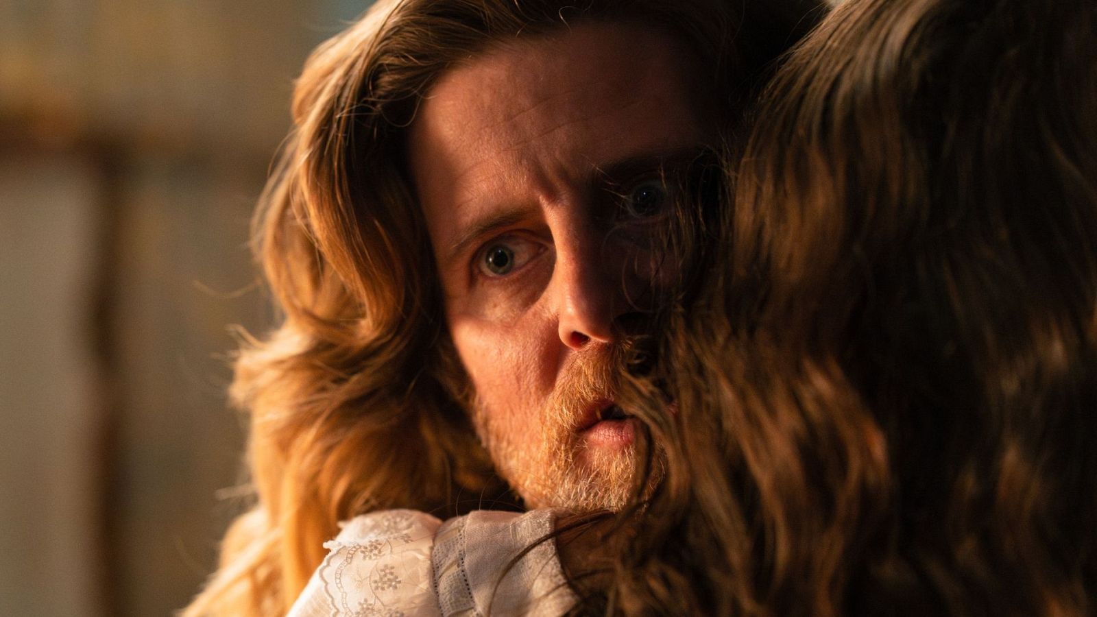 Michael Sheen en primer plano, con ojos muy abiertos y boca entreabierta, mostrando una expresión de shock. Su cabello largo y barba son visibles, junto con el cabello de otra persona que lo rodea.