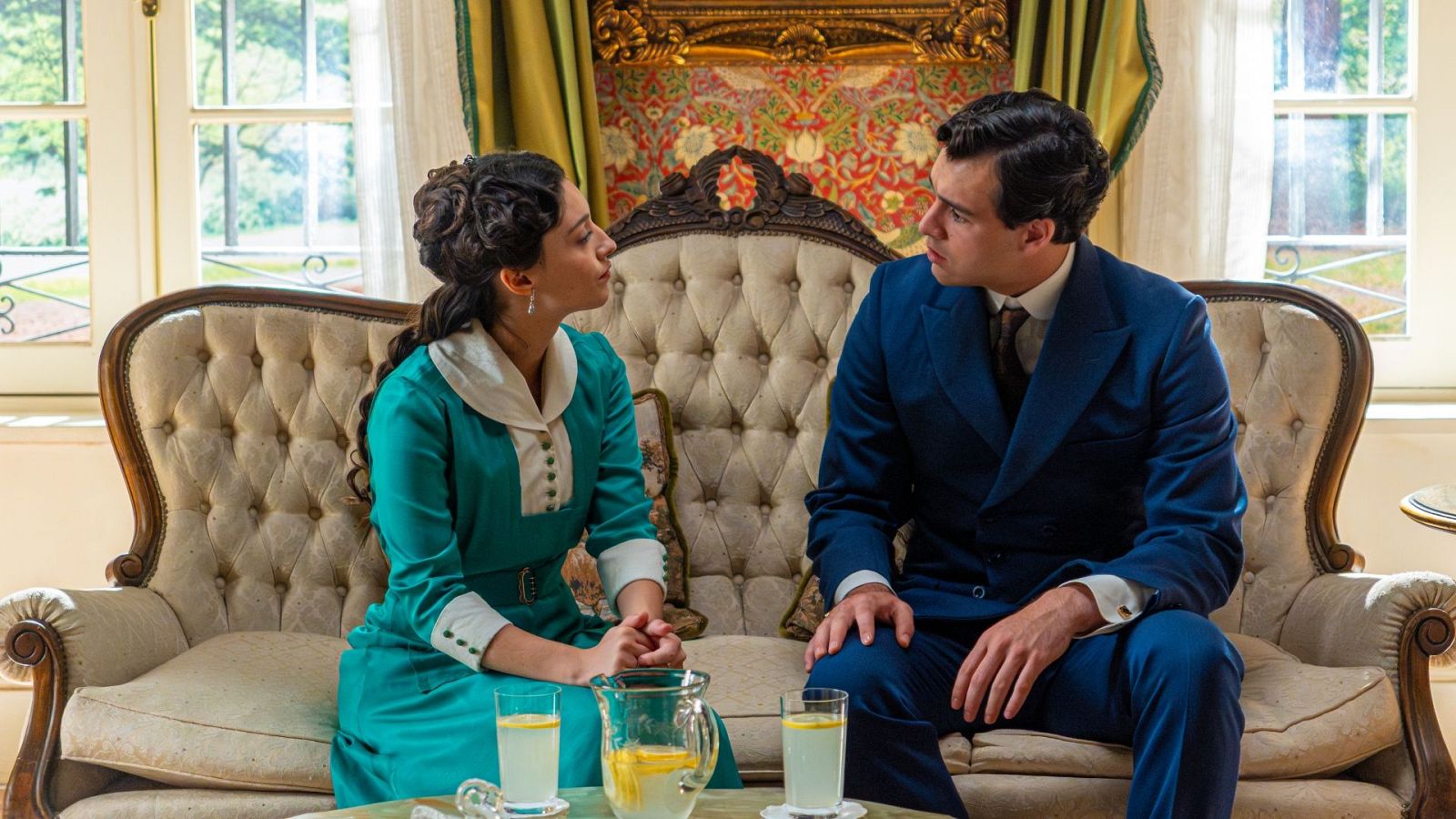 Ana Garcés y Arturo García Sancho, Jana y Manuel de "La Promesa", comparten un momento serio en un sofá clásico, con una mesa de bebidas al frente y un fondo decorado.