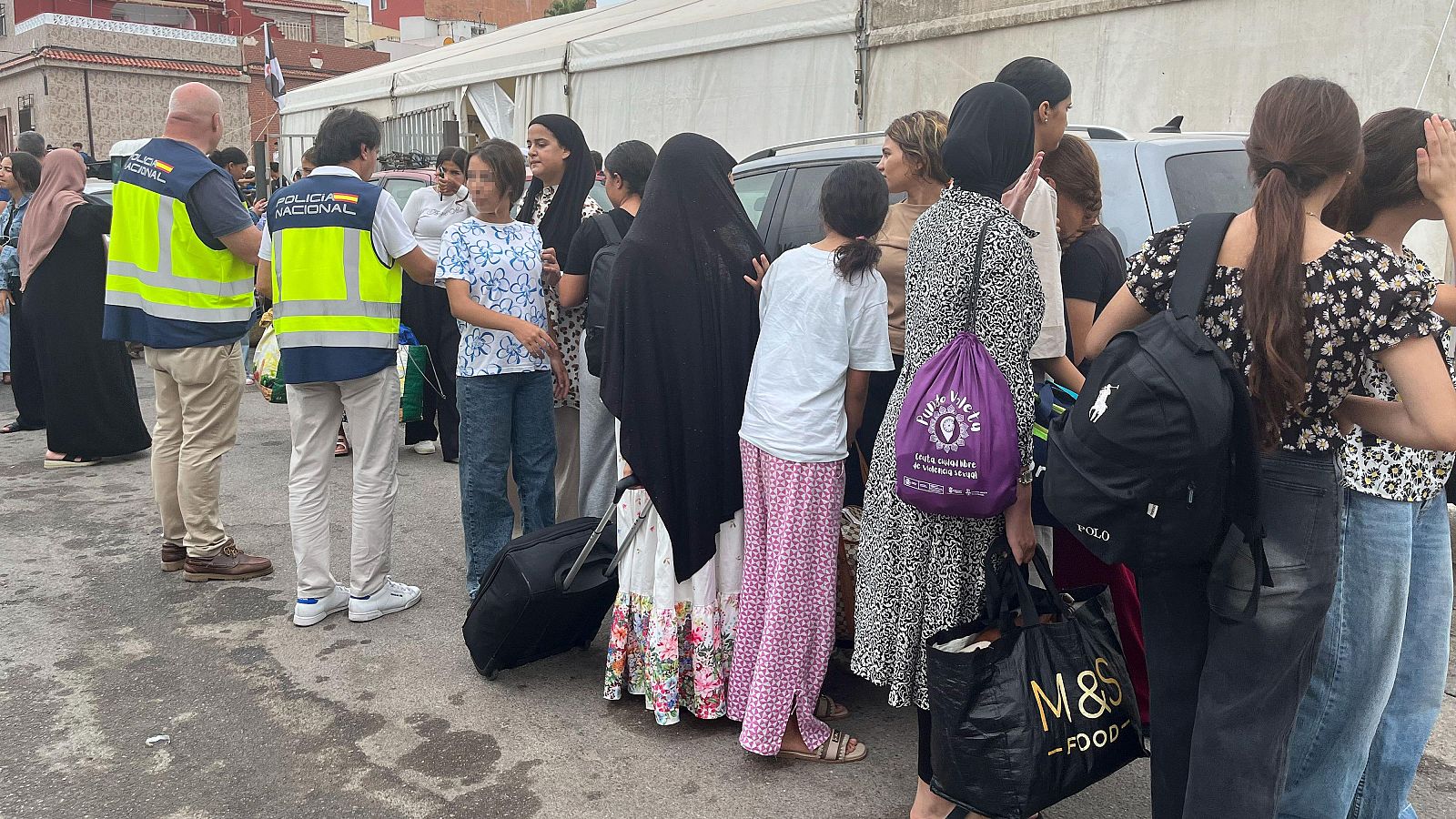 Agentes de Policía Nacional supervisan a un grupo de mujeres y niñas con equipaje en una explanada. Se observan maletas y bolsas de marcas como Polo Ralph Lauren y M&S FOOD.