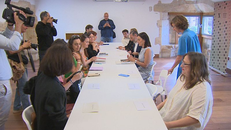 Sala de reuniones con mesa blanca, sillas modernas y asistentes. Equipo de prensa documenta el evento. Fondo con paredes de piedra y lisas, ventanas y soporte con logo.