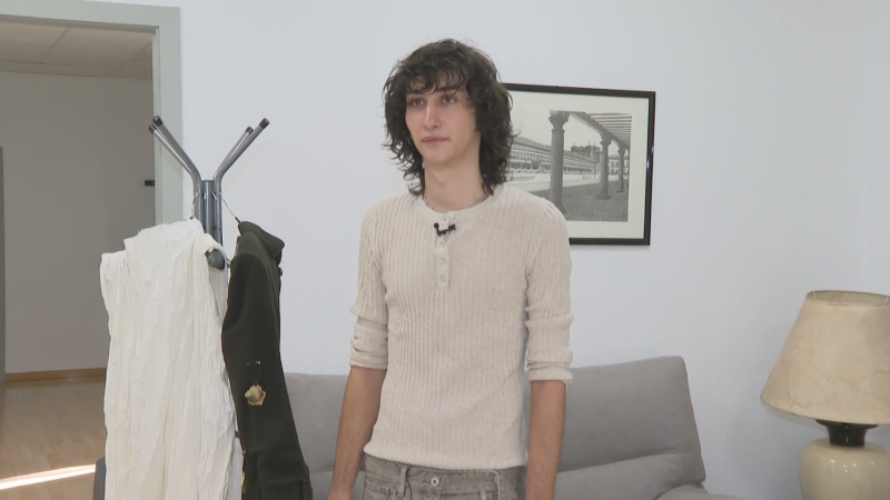 Jorge Juan Selas, con camiseta beige y micrófono, en una sala de entrevistas con perchero y cuadro arquitectónico.