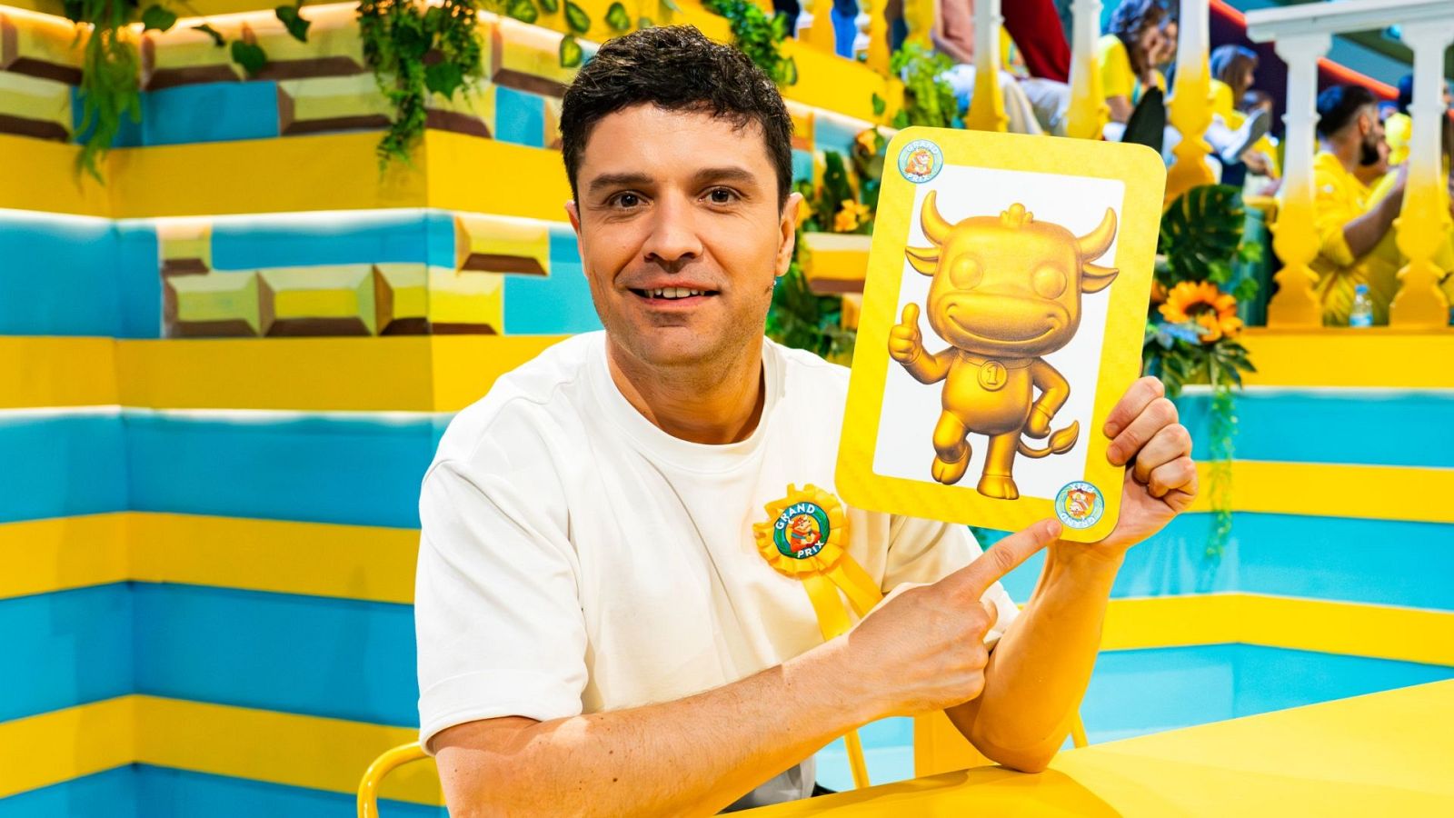 Ramón García sonríe y señala una tarjeta con un toro dorado "pulgar arriba", mascota del "GRAND PRIX", en un estudio colorido.