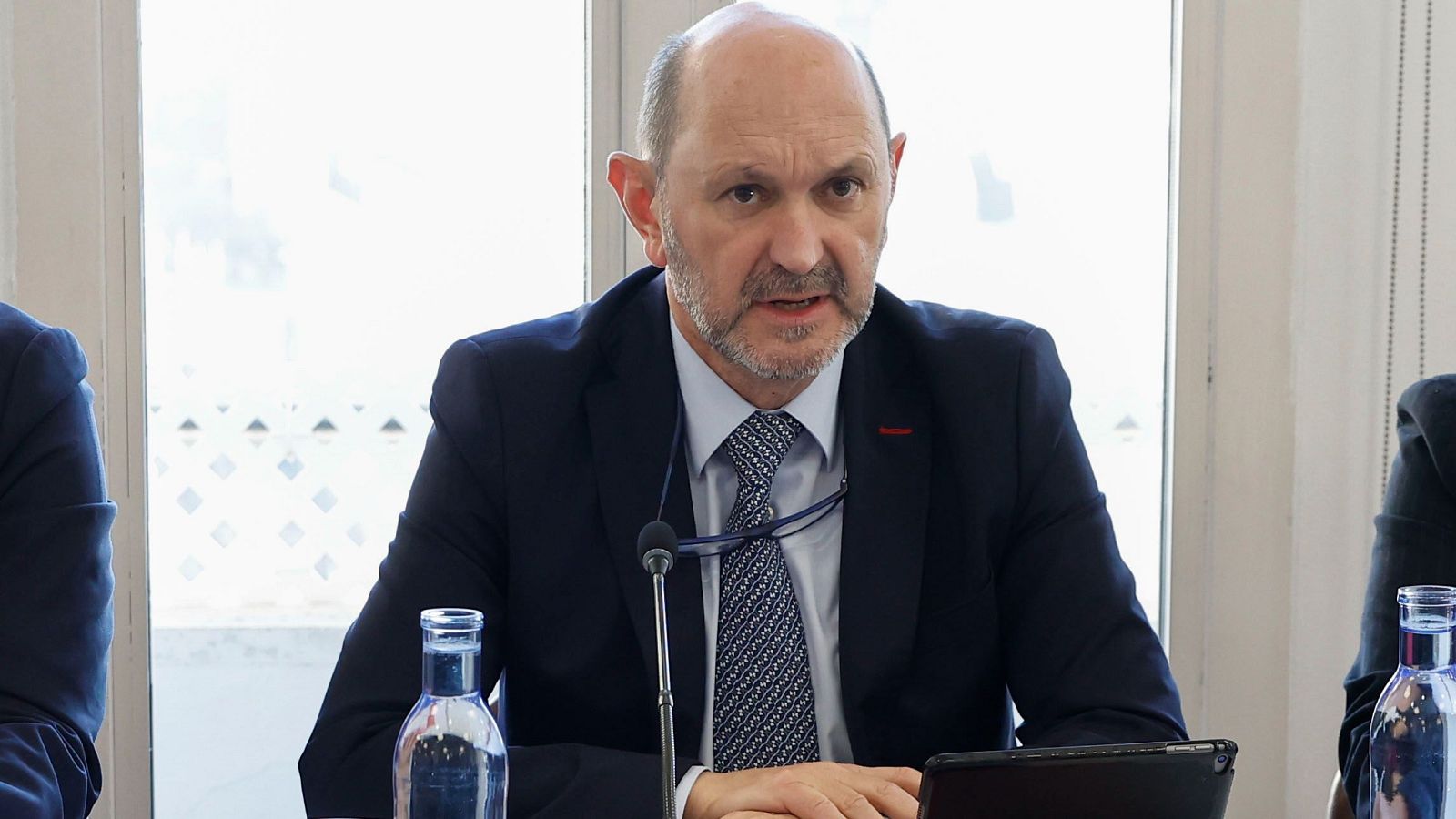 Presidente de la RFEF sentado en mesa con micrófono, tablet y botella de agua.  Fondo con ventana cuadriculada; otras personas presentes.