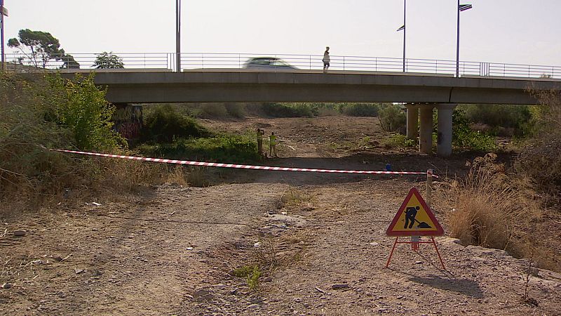Trabajadores en un barranco seco bajo un puente, con señal de "hombres trabajando" y cinta de seguridad.
