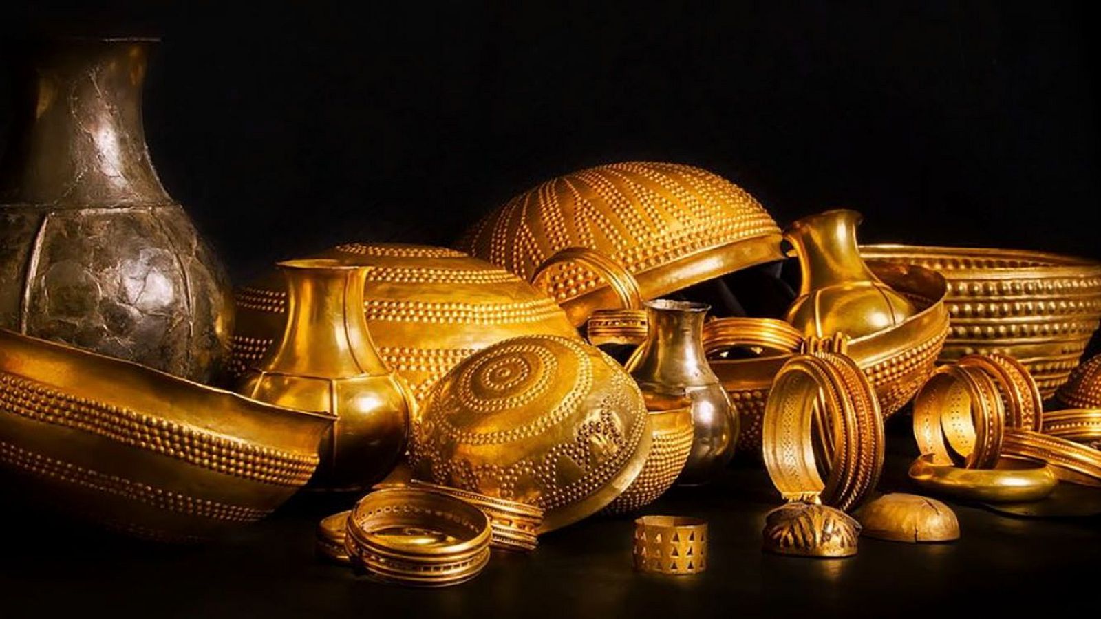 Colección de artefactos antiguos de metal dorado, incluyendo vasijas, cuencos decorados y brazaletes, sobre fondo negro.