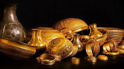 Colecci�n de artefactos antiguos de metal dorado, incluyendo vasijas, cuencos decorados y brazaletes, sobre fondo negro.