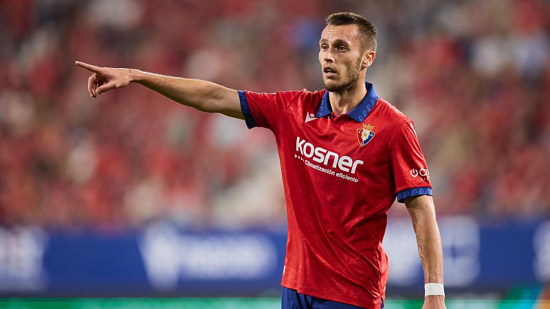 Jonathan Dubasin del CA Osasuna, con camiseta roja y azul, señala con el dedo índice durante un partido de LaLiga EA Sports. Se aprecian el escudo del equipo y el patrocinador "kosner".