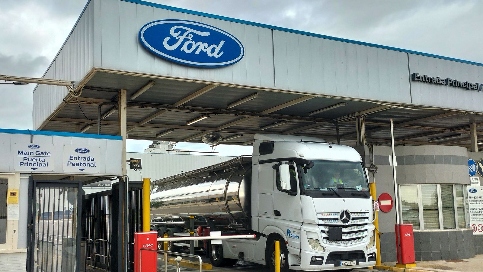 Entrada principal de Ford con camión Mercedes-Benz cisterna detenido, barreras, garita de seguridad y señal de prohibido el paso.