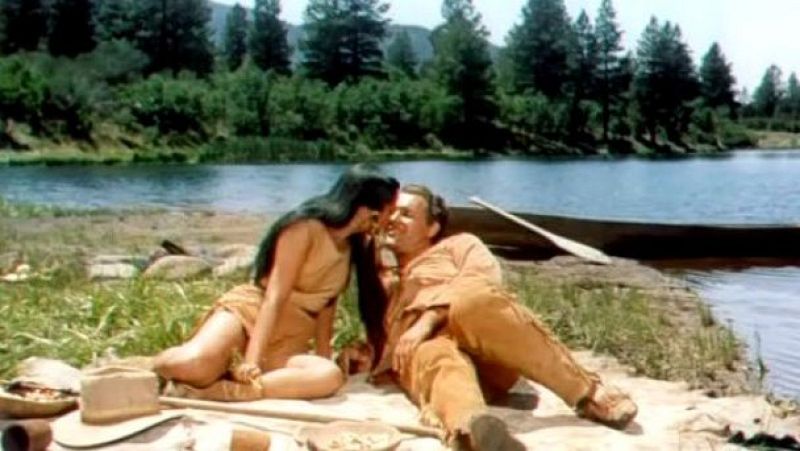 Burt Reynolds y Raquel Welch comparten un momento íntimo al aire libre, con sus rostros casi tocándose, rodeados de naturaleza y una canoa.