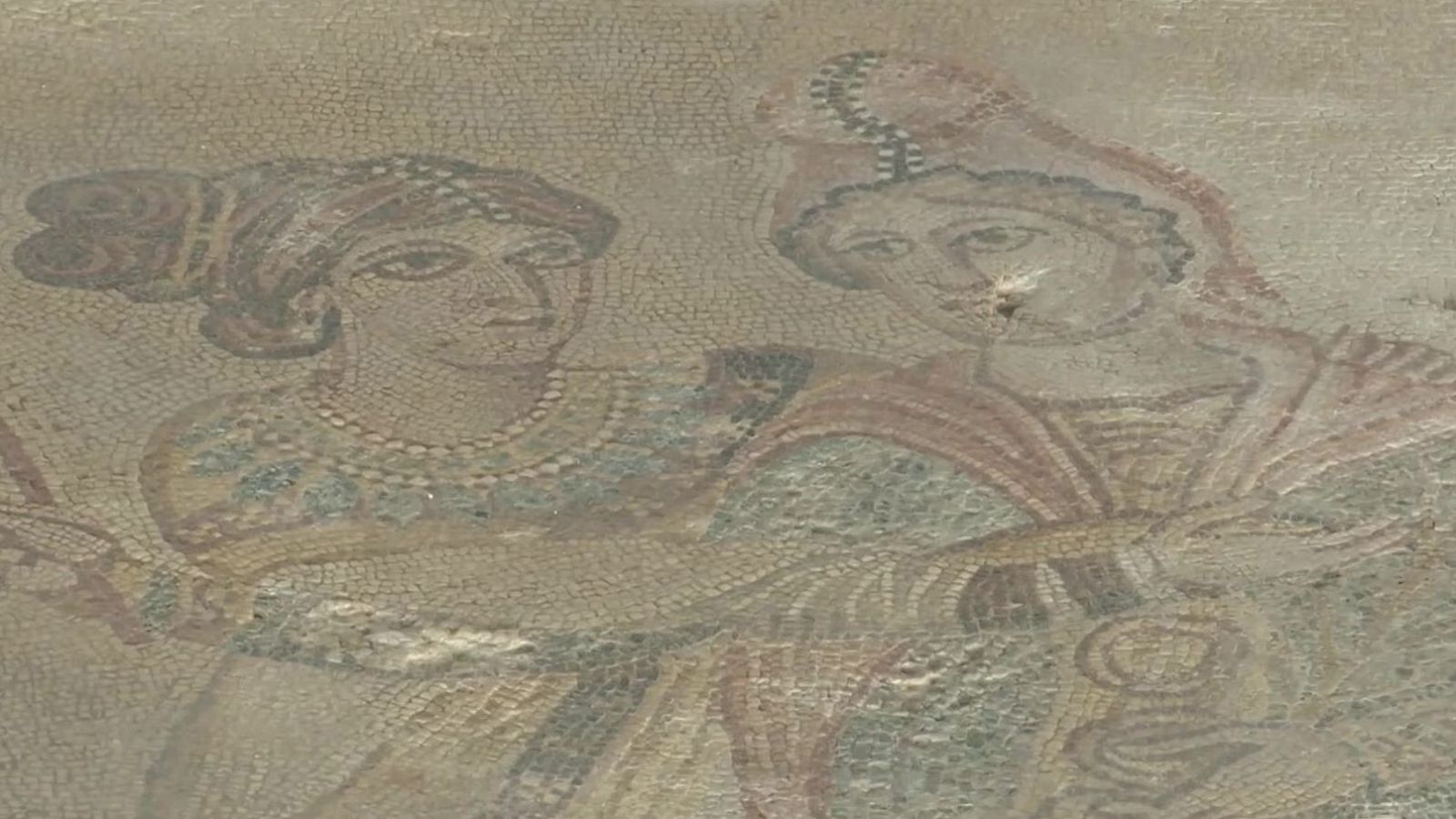 Fragmento de antiguo mosaico con dos figuras femeninas de medio cuerpo, manos unidas al centro. Teselas desgastadas en tonos tierra, ocres, rojizos, azules y verdes apagados.