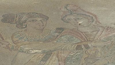 Fragmento de antiguo mosaico con dos figuras femeninas de medio cuerpo, manos unidas al centro. Teselas desgastadas en tonos tierra, ocres, rojizos, azules y verdes apagados.