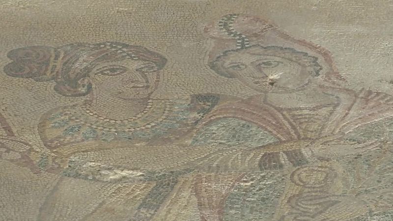 Fragmento de antiguo mosaico con dos figuras femeninas de medio cuerpo, manos unidas al centro. Teselas desgastadas en tonos tierra, ocres, rojizos, azules y verdes apagados.