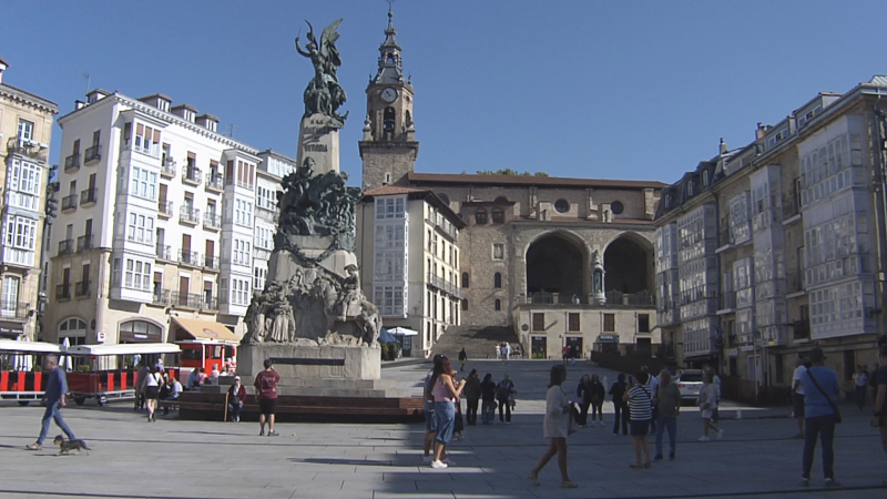 Plaza de la Virgen Blanca, Vitoria-Gasteiz, con el Monumento a la Batalla de Vitoria, Iglesia de San Miguel, gente, edificios y tren turístico bajo cielo azul.