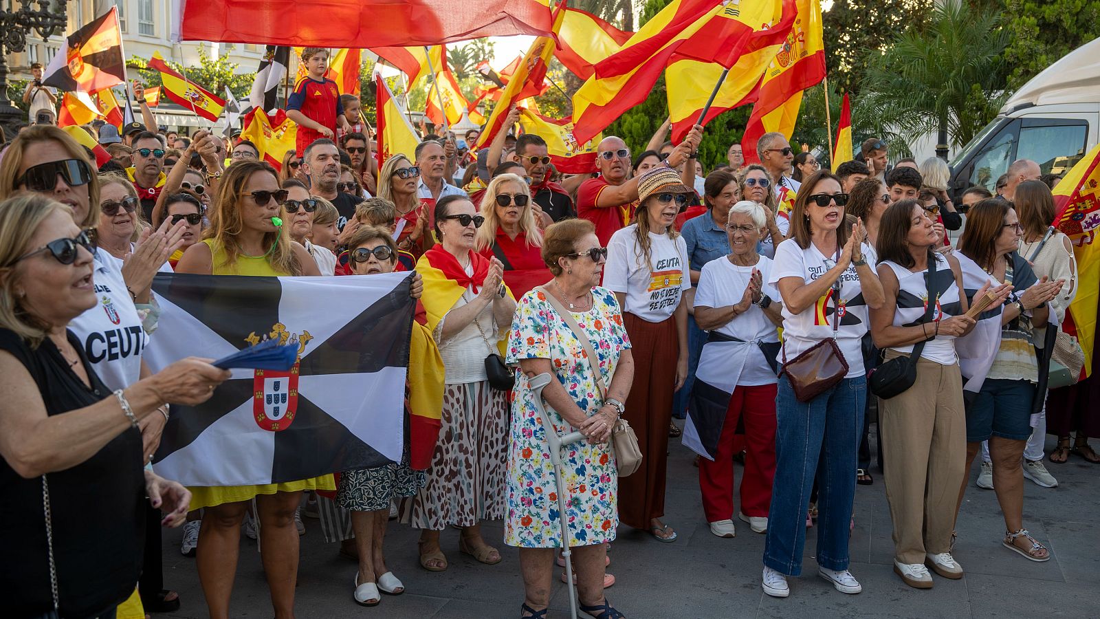 Manifestación pacífica en Ceuta con multitud ondeando banderas de España y Ceuta. Se aprecian mensajes en camisetas y participación activa de adultos y mayores.