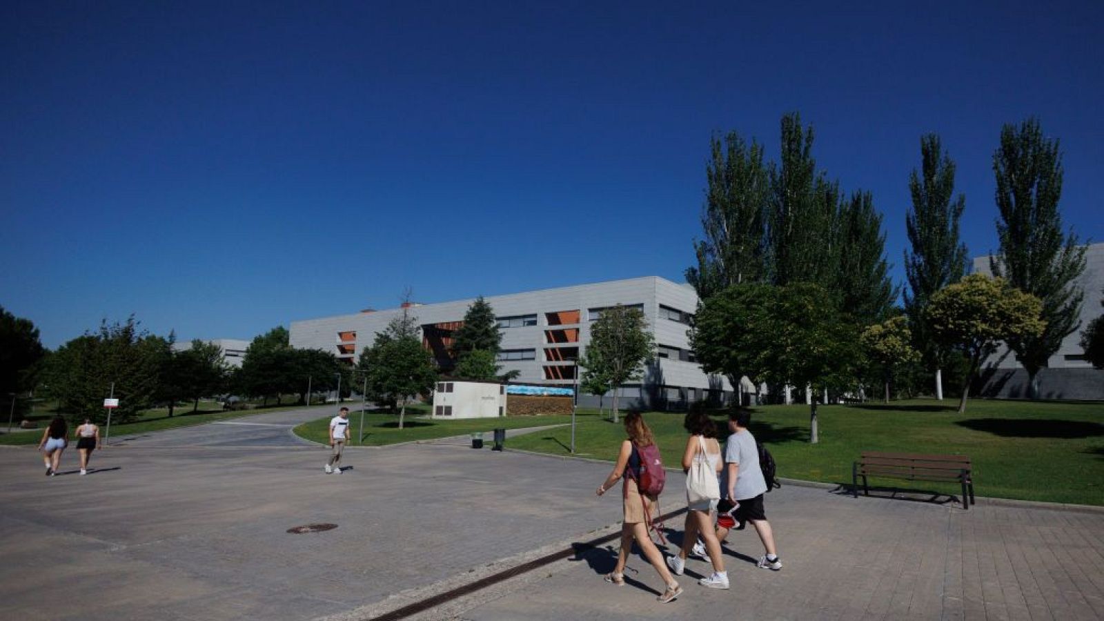 Vista amplia de un campus moderno con edificios grises y terracota, césped, árboles y jóvenes estudiantes caminando por senderos bajo un cielo azul despejado.