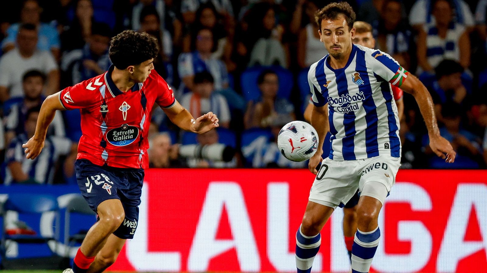 Andrés Antañón (rojo, "Estrella Galicia 0,0") y Mikel Oyarzabal (rayas azules/blancas, "Baghdad Capital", brazalete ikurriña) disputan el balón en un partido de LaLiga.