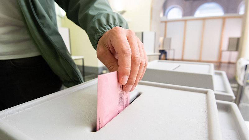 Mano depositando papeleta rosa en urna electoral gris. Se aprecian otras urnas y vestimenta oscura en el fondo.