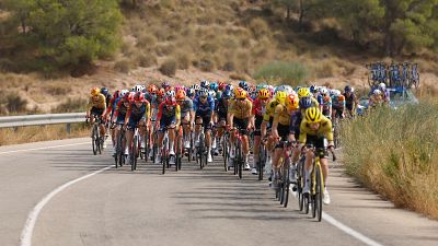 Pelot�n de ciclistas profesionales en carrera por una carretera con colinas y vegetaci�n seca. Equipaciones variadas, cascos, bicicletas de carretera y veh�culo de apoyo visible.