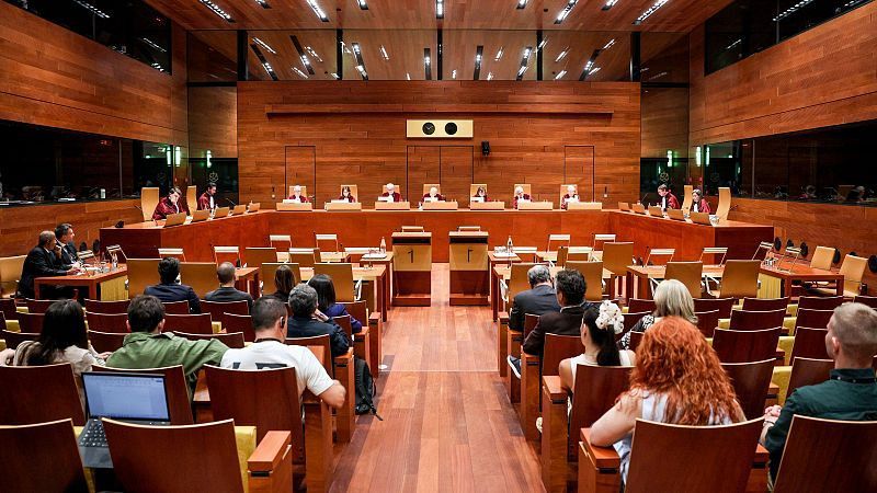 Sala de audiencias moderna con tarima elevada, personas con togas, dos podios con el número "1" y asientos para la audiencia. Predomina la madera cálida en paredes, suelo y mobiliario.