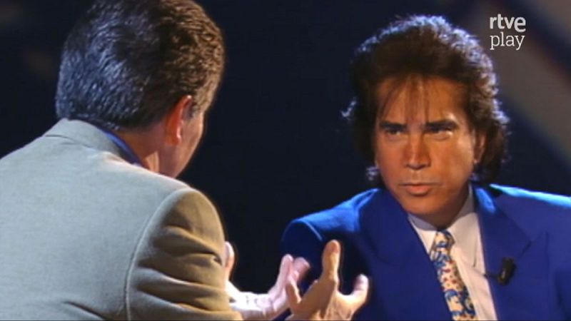 Entrevista a "El Puma" con Pedro Ruiz en "rtve play". El cantante viste traje azul eléctrico, el presentador gesticula. Fondo oscuro de estudio.